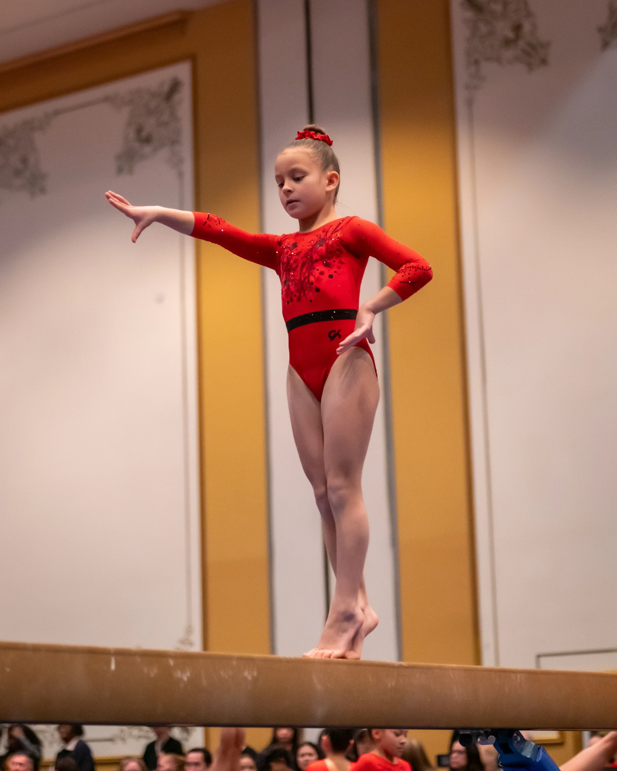A gymnast in a red leotard balances on the beam in the ballroom